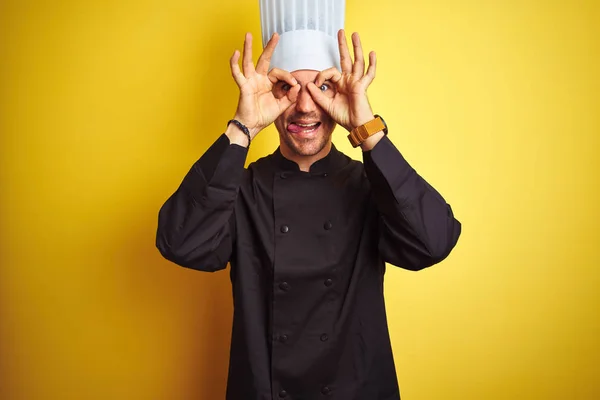 Young chef man wearing uniform and hat standing over isolated yellow background doing ok gesture like binoculars sticking tongue out, eyes looking through fingers. Crazy expression.