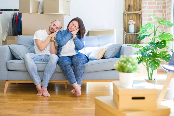 Young couple sitting on the sofa arround cardboard boxes moving to a new house sleeping tired dreaming and posing with hands together while smiling with closed eyes.