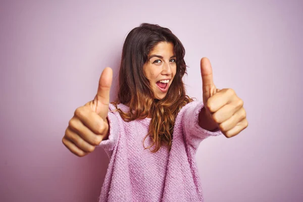 Young beautiful woman wearing sweater standing over pink isolated background approving doing positive gesture with hand, thumbs up smiling and happy for success. Winner gesture.
