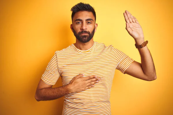 Young indian man wearing t-shirt standing over isolated yellow background Swearing with hand on chest and open palm, making a loyalty promise oath