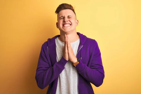 紫色のスウェットシャツを着た若いハンサムな男性は 孤立した黄色の背景の物乞いをして手で祈り 顔には希望の表現と一緒に非常に感情的で心配して立っている 許しを求める 宗教概念 — ストック写真