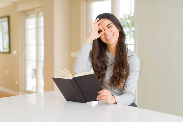Young woman reading a book with happy face smiling doing ok sign with hand on eye looking through fingers
