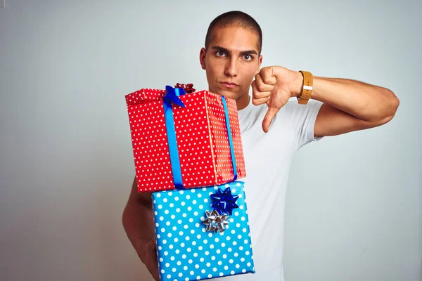 Young handsome man holding birthday gifts over white isolated background with angry face, negative sign showing dislike with thumbs down, rejection concept