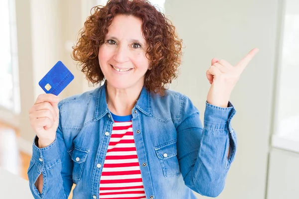 Senior woman holding credit card as payment very happy pointing with hand and finger to the side
