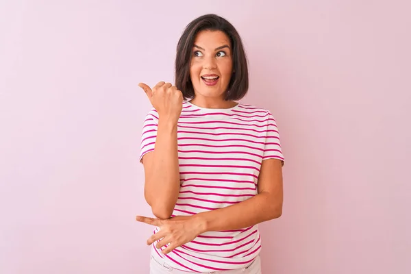 年轻的美丽女人穿着条纹T恤站在孤立的粉红色背景微笑与幸福的脸看 并指着一边竖起大拇指 — 图库照片