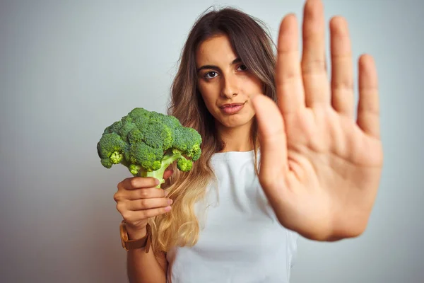 若いです美しいです女性食べるブロッコリー以上グレー隔離された背景とともにオープン手Doing停止記号で深刻で自信を持って表現 防衛ジェスチャー — ストック写真