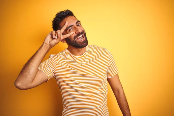 孤立した黄色の背景の上に立つTシャツを着た若いインド人男性が顔の上に指で平和のシンボルを行い 勝利を示す陽気な笑顔 — ストック写真