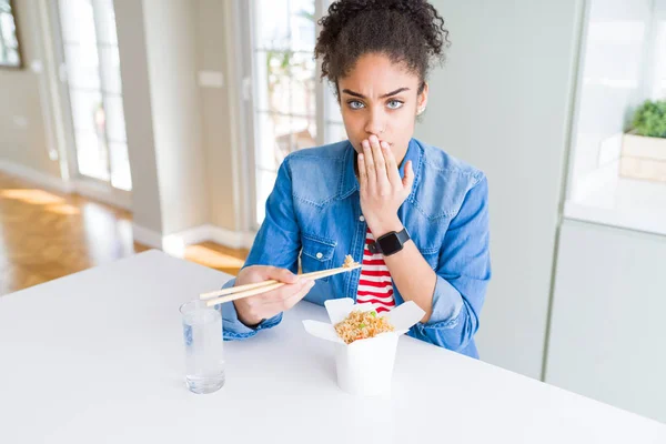 Young african american woman eating asian noodles from delivery box cover mouth with hand shocked with shame for mistake, expression of fear, scared in silence, secret concept