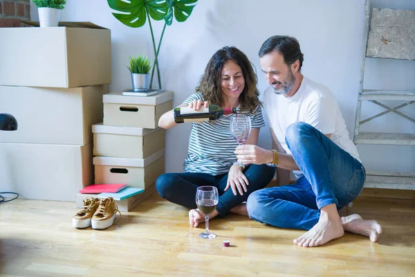 Orta yaş Senior romantik çift aşk yeni bir eve taşımak için mutlu gülümseyen bir bardak şarap içme kutluyor, etrafında kutuları ile daire katında oturan