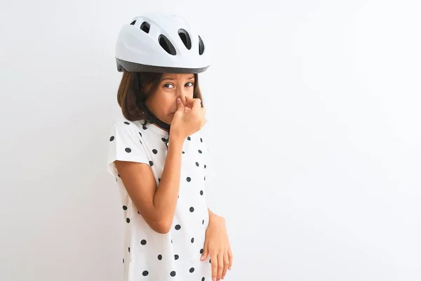 Beautiful child girl wearing security bike helmet standing over isolated white background smelling something stinky and disgusting, intolerable smell, holding breath with fingers on nose. Bad smells concept.
