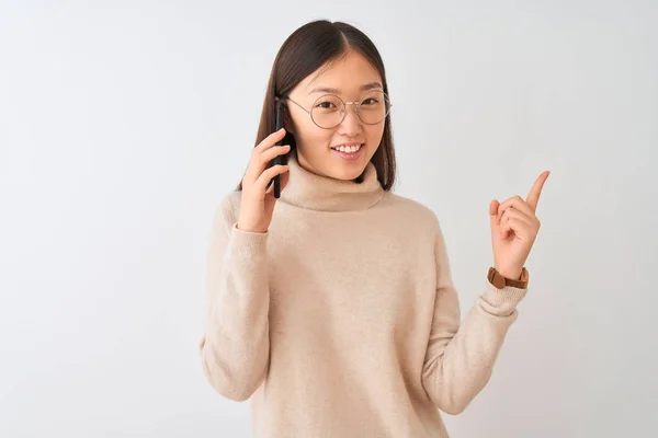 顔に大きな笑顔で隔離された白い背景の上にスマートフォンで話している若い中国人女性 カメラを見て側に手指で指して — ストック写真