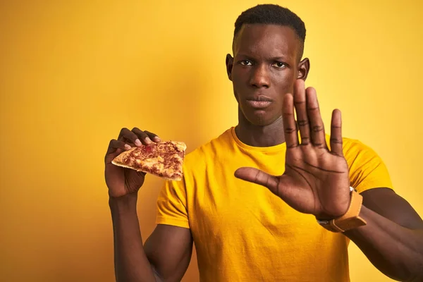 African american man eating slice of pizza standing over isolated yellow background with open hand doing stop sign with serious and confident expression, defense gesture