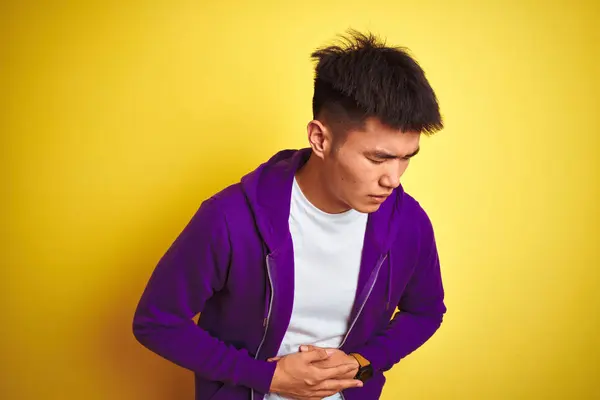 消化不良 痛みを伴う病気が体調不良のため 若いアジアの中国人男性が胃の上に手で孤立した黄色の背景の上に立って紫色のスウェットシャツを着て アチェの概念 — ストック写真