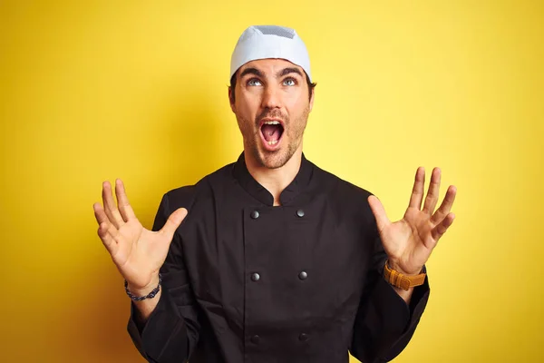 年轻英俊的厨师 穿着制服和帽子在孤立的黄色背景上做饭 疯狂而疯狂地大喊大叫 带着咄咄逼人的表情和手臂 沮丧的概念 — 图库照片