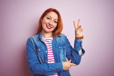 Güzel kızıl saçlı kadın kot gömlek ve pembe arka planda çizgili tişört giyerek kameraya gülümsüyor ve parmaklarıyla zafer işareti yapıyor. İki numara..