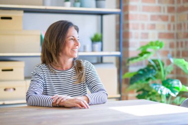 Orta yaş yaşlı kadın evde masada oturan yüzünde gülümseme ile yan bakıyor, doğal ifade. Gülmekten emin.