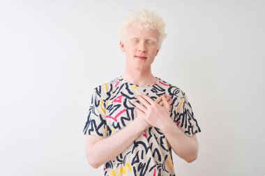 Renkli tişört giyen genç albino bir adam izole edilmiş beyaz arka plan üzerinde duruyor elleri göğsünde, gözleri kapalı ve yüzünde minnet dolu bir jest ile gülümsüyor. Sağlık kavramı.