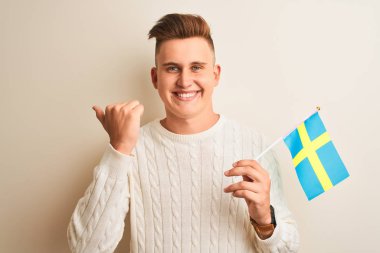 İsveçli genç adam İsveç bayrağını izole edilmiş beyaz arka plan üzerinde tutuyor ve baş parmağıyla gülümsüyor.