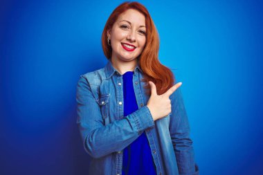 Genç, güzel, kızıl saçlı, kot gömlek giyen, mavi izole edilmiş arka planda neşeli, yüzünde gülümseme, mutlu ve doğal bir ifadeyle elini ve parmağını yana gösteren bir kadın.