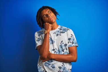 Rasta desenli tişörtlü Afro-Amerikan bir adam izole edilmiş mavi arka plan üzerinde elleri çenesinde soru sorarken düşünceli bir ifade takınıyor. Düşünceli bir yüzle gülümsüyor. Şüphe kavramı.