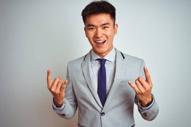 Asyalı Çinli işadamı gri ceketli ve kravatlı, izole edilmiş beyaz arka planda duruyor çılgın bir ifadeyle elleri havada rock sembolü yapıyor. Müzik yıldızı. Ağır konsept.