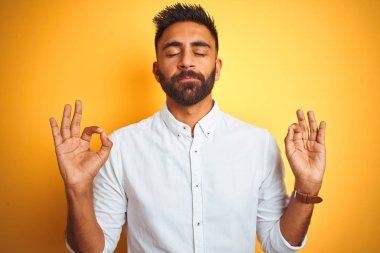İzole edilmiş sarı arka plan üzerinde duran beyaz şık gömlekli Hintli işadamı rahatla ve parmaklarıyla meditasyon yaparken gözleri kapalı gülümse. Yoga konsepti.