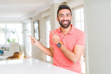 Evde rahat t-shirt giyen İspanyol yakışıklı gülümseyen ve iki elleri ve parmakları ile tarafına işaret kamera bakıyor.