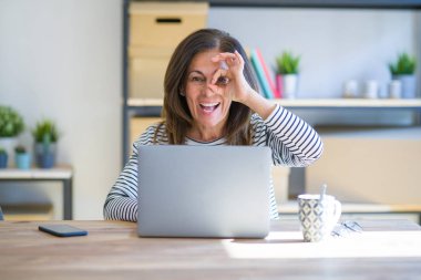 Orta yaşlı bir kadın evinde oturmuş bilgisayarla çalışıyor. El hareketleriyle gülümsüyor ve mutlu yüzüyle parmaklarının arasından bakıyor..