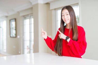 Güzel Asyalı kadın beyaz masada kırmızı kazak giyiyor gülümsüyor ve kameraya bakıyor iki eliyle ve parmaklarıyla kenarı işaret ediyor..