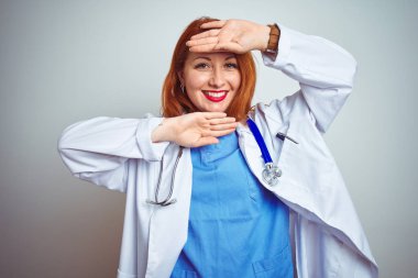 Beyaz izole arka plan üzerinde stetoskop kullanarak genç Kızıl saçlı doktor kadın gülümseyen neşeli oyun Peek elleri ile yüz gösteren bir Boo. Sürpriz ve çıkıldı