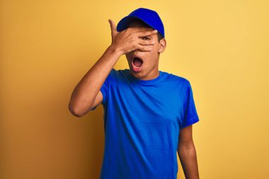 Yakışıklı Arap teslimatçı, izole edilmiş sarı arka planda duruyor. Şoka bakıyor. Yüzü ve gözleri kapanıyor, utangaç ifadesiyle parmaklarından bakıyor..