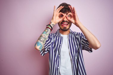 İzole edilmiş pembe arka plan üzerinde duran çizgili tişörtlü genç adam dürbün gibi güzel hareketler yapıyor. Dürbün dilini dışarı çıkarıyor, gözler parmaklarından bakıyor. Çılgın ifade.