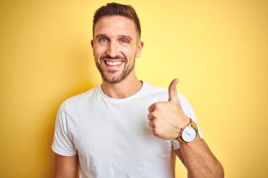 Sarı izole edilmiş arka plan üzerine gündelik beyaz tişört giyen genç yakışıklı adam mutlu bir el hareketi yapıyor. Kameraya bakıp başarıyı gösteren ifade onaylanıyor.