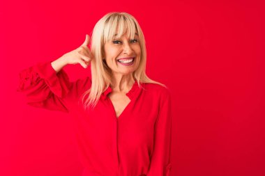 Soyutlanmış kırmızı arka planda duran zarif gömlekli, orta yaşlı bir kadın telefonla el ve parmaklarıyla telefonla konuşuyor gibi gülümsüyor. İletişim kavramları.