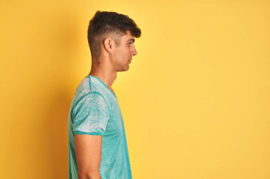 Yeşil t-shirt giyen genç Hintli adam izole edilmiş sarı arka planda duruyor, yan yana bakıyor, rahat profil pozu veriyor, kendine güveni tam bir gülümsemeyle..