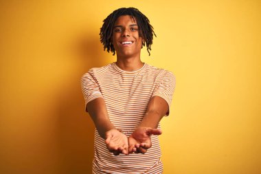 Rastalı tişörtlü Afro bir adam izole edilmiş sarı arka planda ayakta duruyor avuç içleriyle gülümsüyor ya da jest yapıyor. Tutun ve koruyun