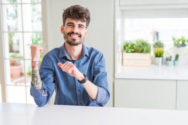 Genç adam beyaz bir masada oturuyor. Arka tarafı işaret ediyor. El ve baş parmakları havada, kendinden emin bir şekilde.