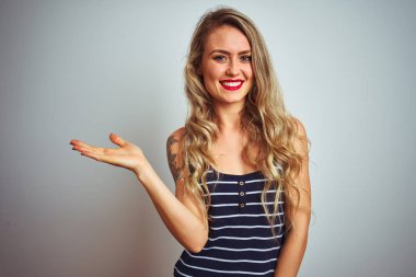 Çizgili tişört giyen genç bir kadın beyaz izole edilmiş arka planda dikiliyor neşeli bir şekilde gülümsüyor ve avuç içiyle kameraya bakıyor..