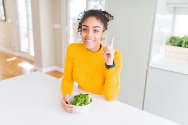 Genç Afrikalı Amerikalı kız sıcak yeşil brokoli yiyor. Bir fikir ya da soruyla şaşırmış. Mutlu bir yüzle işaret ediyor.