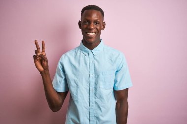Mavi gömlekli Afro-Amerikalı bir adam izole edilmiş pembe arka planda dikiliyor. Kameraya gülümsüyor ve zafer işareti yapıyor. İki numara..