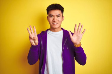 Mor kazak giyen Asyalı genç adam izole edilmiş sarı arka planda duruyor ve 8 numaralı parmaklarıyla işaret ederken kendinden emin ve mutlu bir şekilde gülümsüyor..