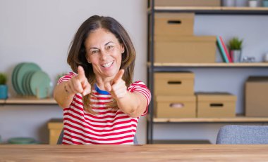 Orta yaş yaşlı kadın evde masada oturan ve parmakları ile kamera işaret, olumlu ve neşeli gülümseyen