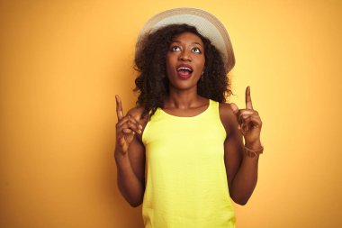 Genç Afro-Amerikalı kadın tişört ve şapka giyip izole edilmiş sarı arka planda şaşkın şaşkın bakıyor ve parmaklarıyla işaret ediyor ve kollarını kaldırıyordu..