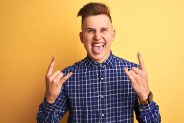 Genç yakışıklı adam rahat gömlek giyen izole sarı arka plan üzerinde duran deli ifade elleri ile rock sembolü yapıyor bağırarak. Müzik yıldızı. Ağır konsept.