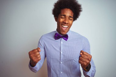 İzole edilmiş beyaz arka plan üzerinde çizgili gömlek ve mor papyon takan afro iş adamı çok mutlu ve heyecanlı bir şekilde kollarını kaldırıp, gülümseyerek ve başarı için çığlık atarak zafer hareketi yapıyor. Kutlama konsepti.
