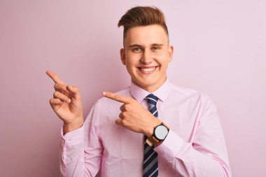 Genç yakışıklı iş adamı gömleği ve kravatıyla pembe arka planda dikiliyor gülümsüyor ve kameraya bakıyor iki eliyle ve parmaklarıyla yan tarafa bakıyor..