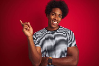 Afro saçlı genç Amerikalı adam izole edilmiş kırmızı arka plan üzerinde deniz mavisi çizgili tişört giyiyor. Yüzünde kocaman bir gülümseme var. Kameraya el ve parmak ile bakıyor..