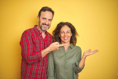 Izole sarı arka plan üzerinde Güzel orta yaş çift şaşırttı ve el ile sunarken kameraya gülümseyerek ve parmak ile işaret.
