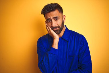 Sarı arka planda üniforma giyen yakışıklı Hintli işçi çapraz kollu depresyondan sıkılmış ve yorgun görünüyor..