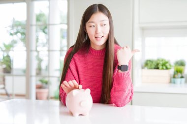 Güzel Asyalı kadın domuz çantasına bozuk para koyuyor ve gülümseyen bir gülümsemeyle baş parmağıyla işaret ediyor.